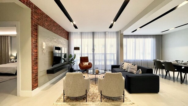 A modern living room with a brick accent wall, a flat-screen TV, a sofa, two armchairs, a coffee table, and a dining area with a table and chairs. - C Living - Premium 3 Bedroom - AQIQ (Riyadh)