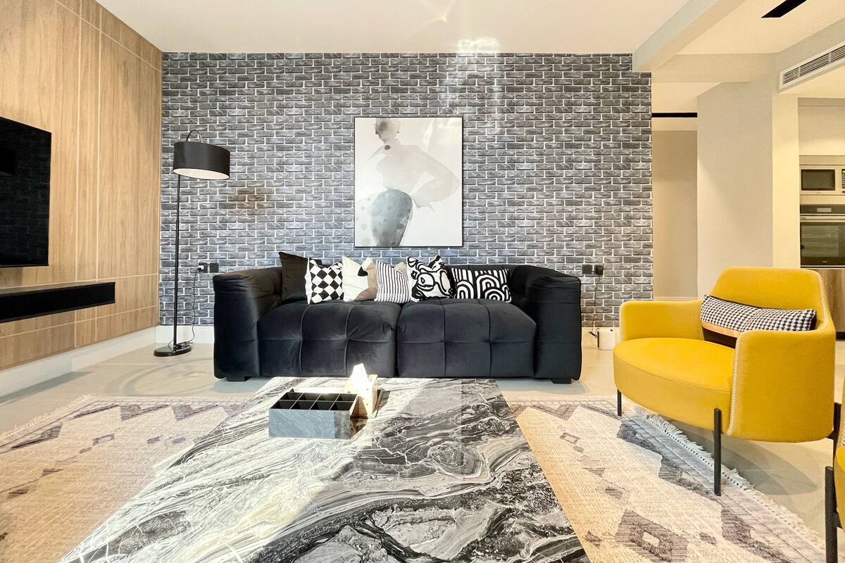 A modern living room with a black leather sofa, a yellow armchair, a marble coffee table, and a brick-patterned wall.