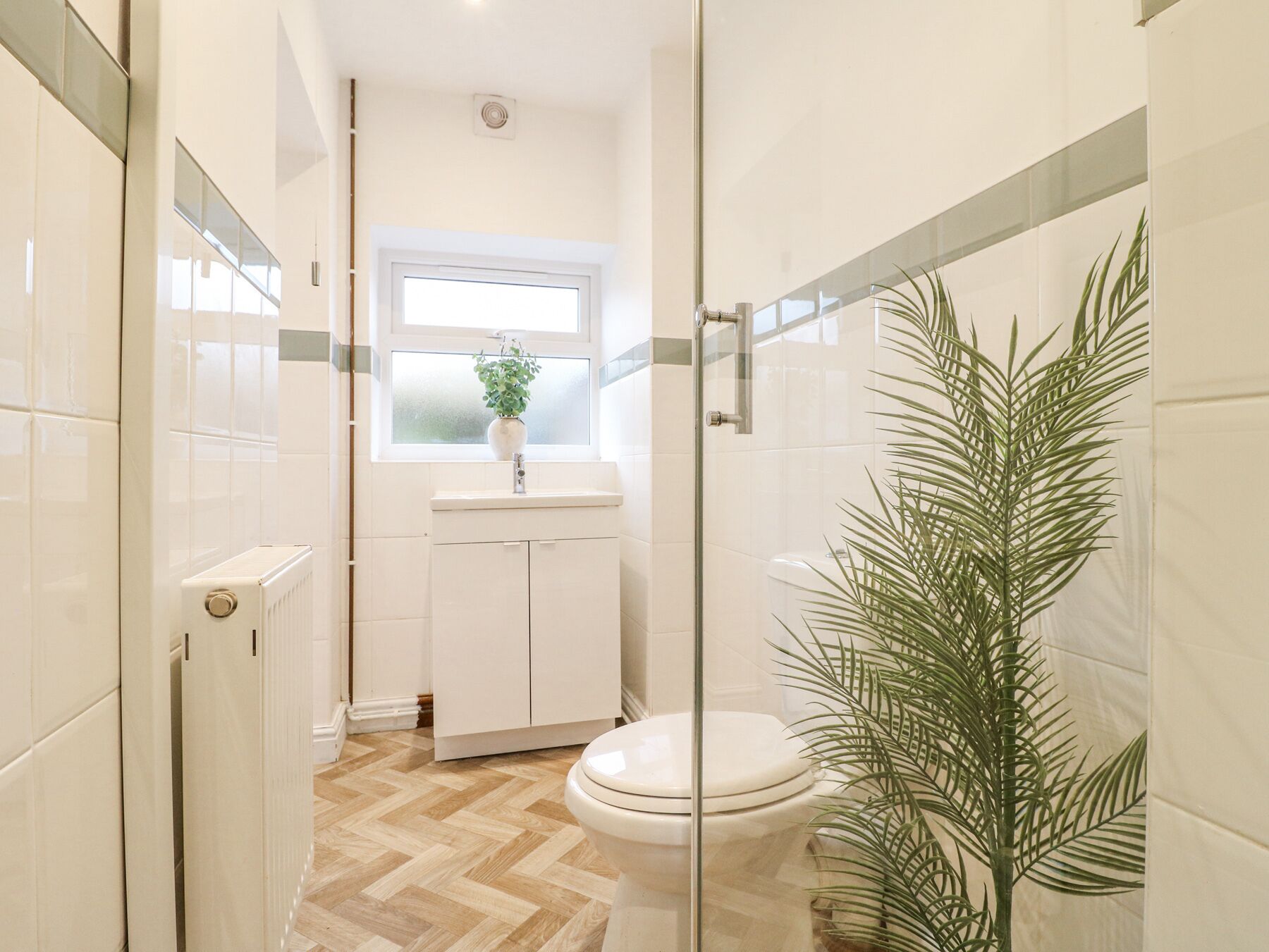 A modern bathroom with a glass shower enclosure, a white toilet, a small window with a plant, and a radiator.