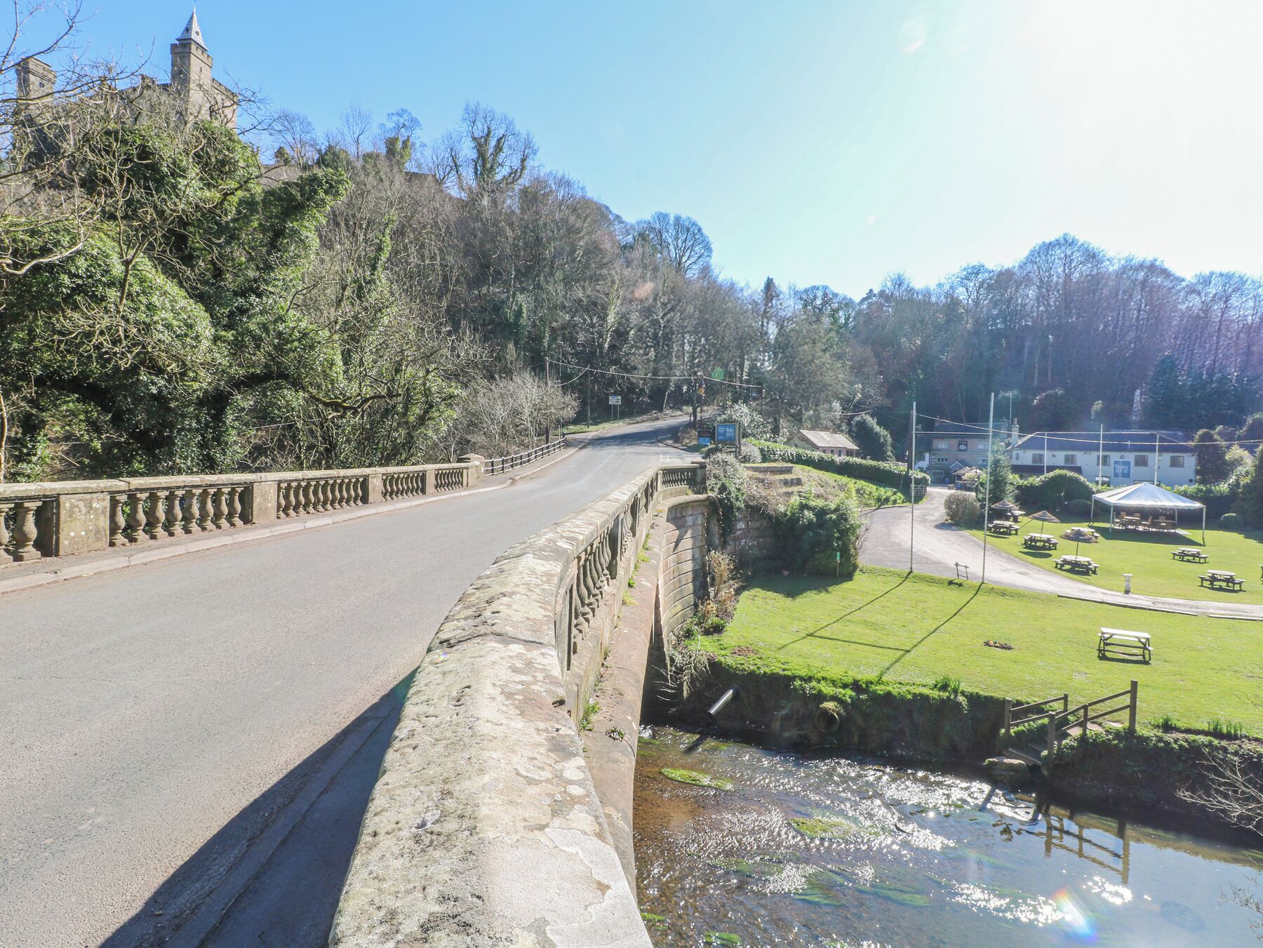 Un pont de pierre enjambant une rivière, une zone herbeuse avec des tables de pique-nique et un bâtiment en arrière-plan.