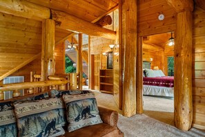 A log cabin interior with a wooden staircase, a cozy seating area with animal-themed cushions, and a bedroom with a bed and a window.