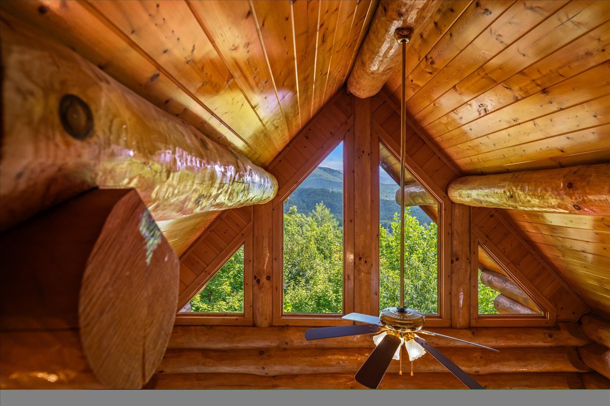 A wooden cabin with a sloped roof, a ceiling fan, and a view of green trees through a window.