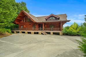 A large log cabin with a spacious front porch, stone foundation, and a clear driveway.