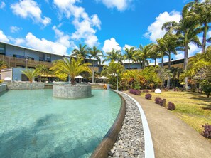 A swimming pool with a stone edge, surrounded by palm trees and a grassy area with a building in the background. - Be Cosy Apart Hotel with LOV (Trou aux Biches)
