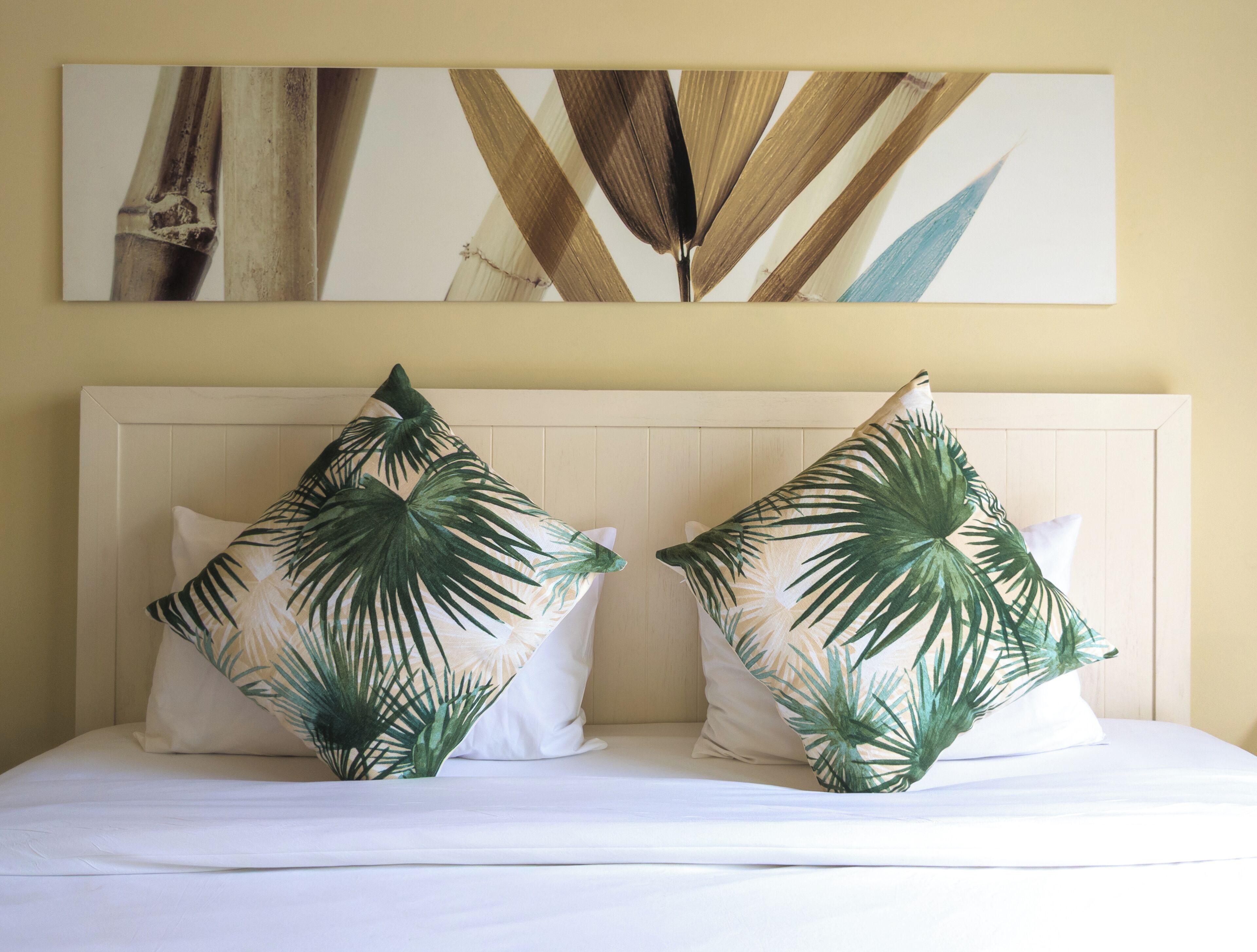 A bed with two pillows featuring palm leaf patterns, a white headboard, and a wall with abstract art.