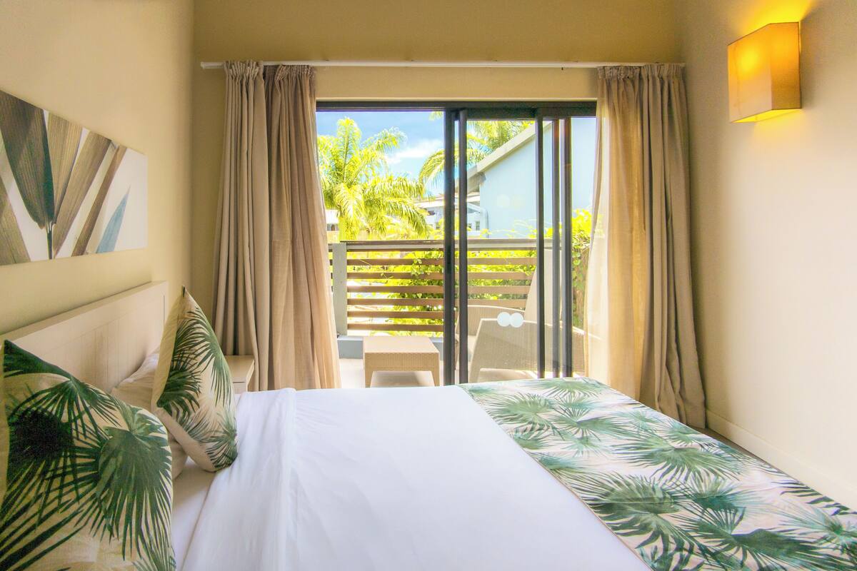 Habitación de hotel con cama, cortinas, vista a un balcón con sillas y una colcha con estampado de hojas de palmera.