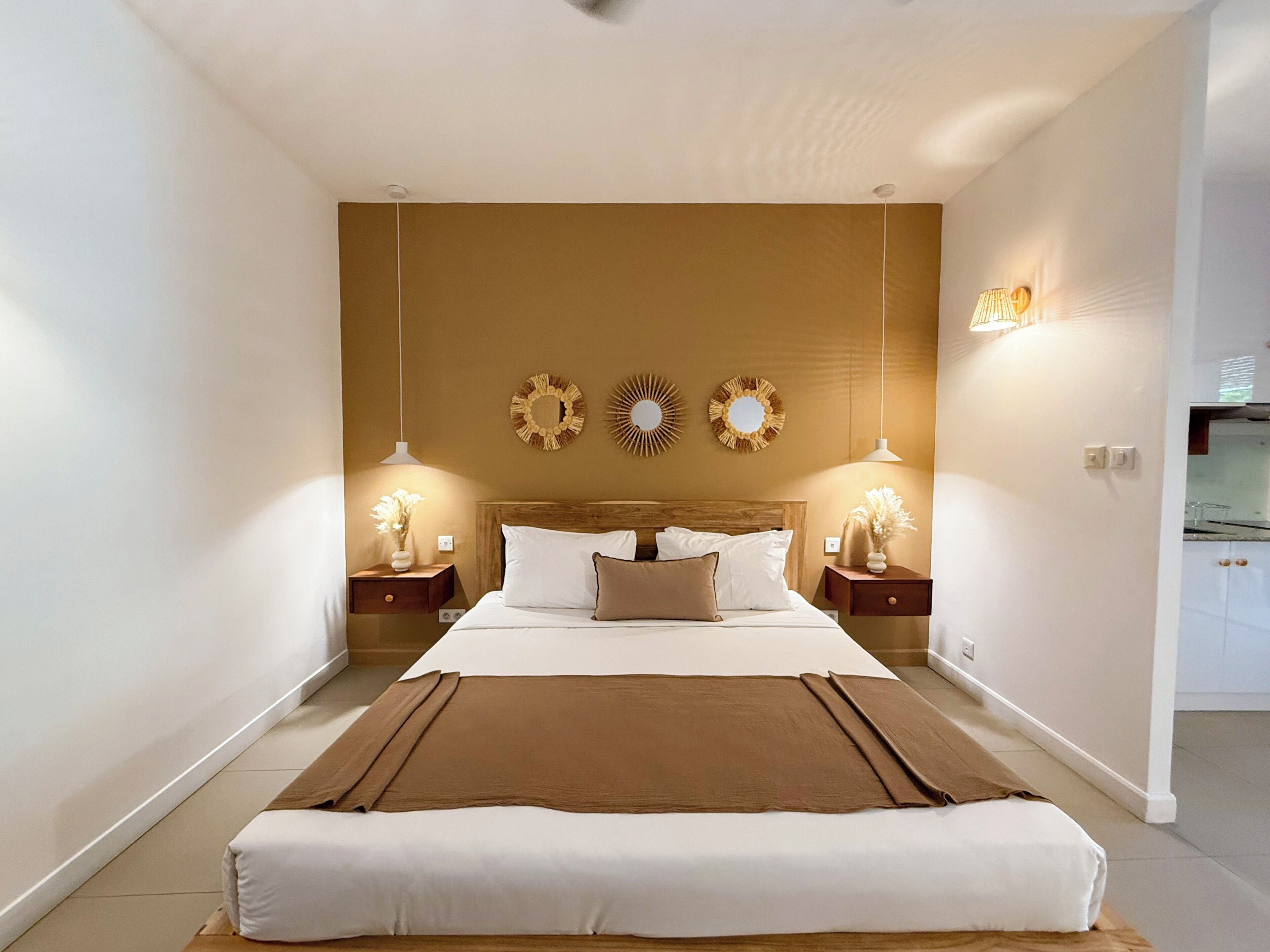 A hotel room with a large bed, two bedside tables, a nightstand, a wall-mounted mirror, and a small kitchen area.