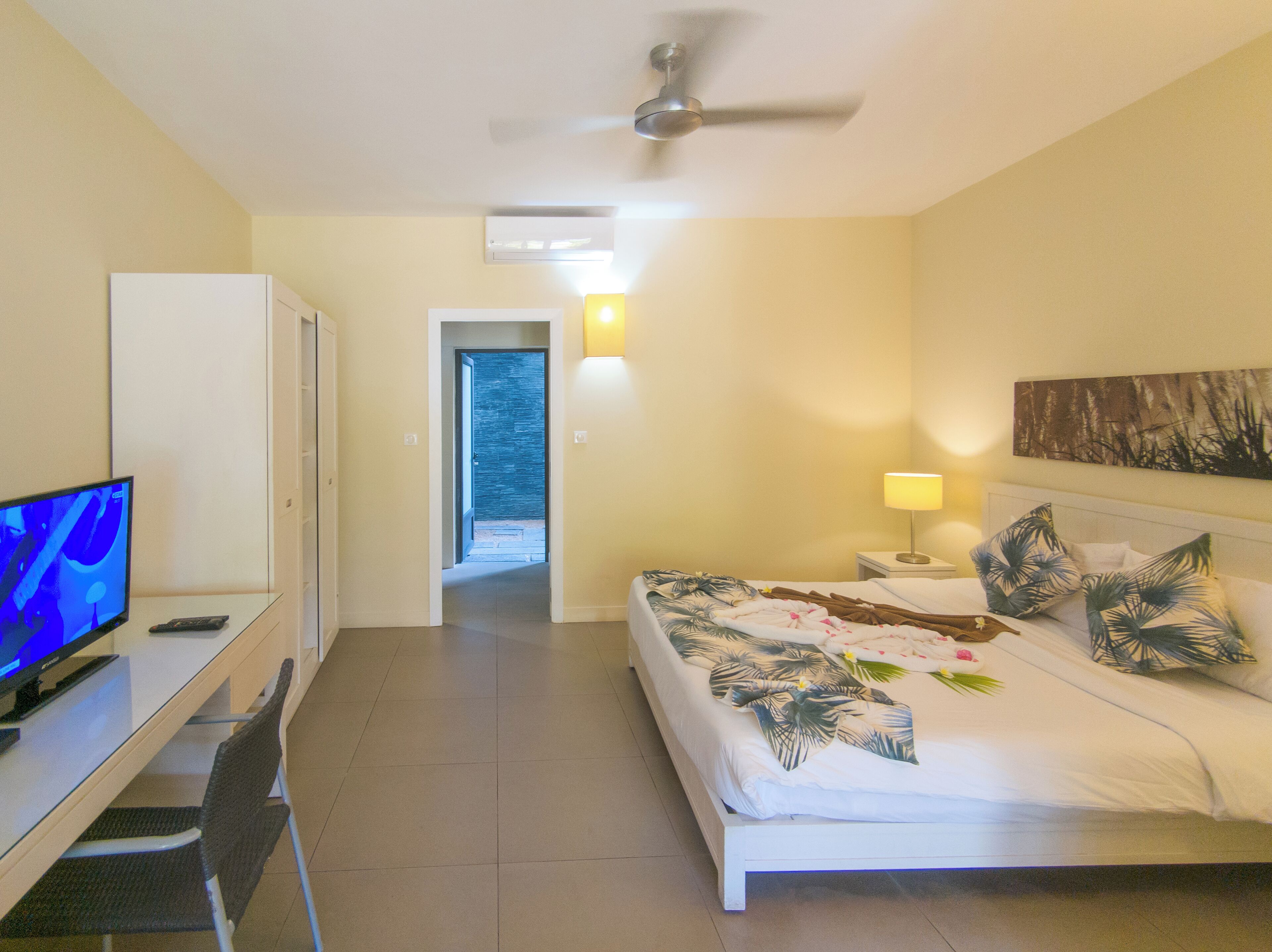 Habitación de hotel con cama, escritorio con computadora, televisor, ventilador de techo y puerta que da a un balcón.