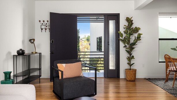 A modern living room with a dark sofa, a small table, a potted plant, and a view through an open door to the outside.