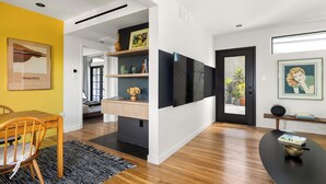 A modern living room with a wooden dining table, a black sofa, and a TV mounted on the wall.