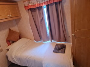 Um quarto pequeno e acolhedor, com uma cama de solteiro, uma janela com cortinas, um guarda-roupa de madeira e uma porta.