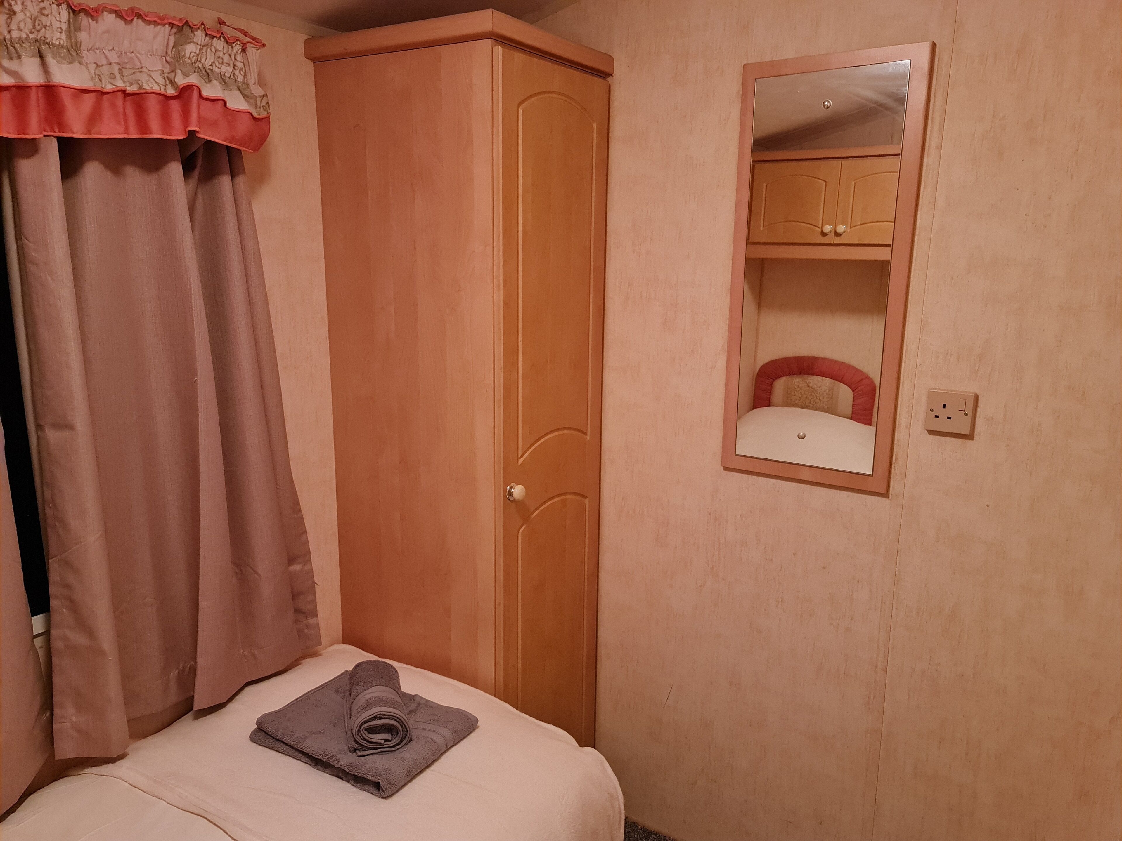 A bedroom with a wooden wardrobe, a bed with a rolled-up towel, a mirror, and a wall socket.