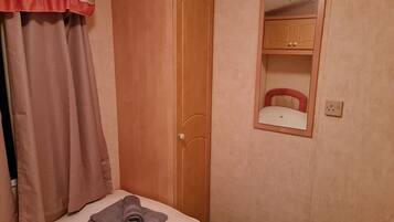 A bedroom with a wooden wardrobe, a bed with a rolled-up towel, a mirror, and a wall socket.
