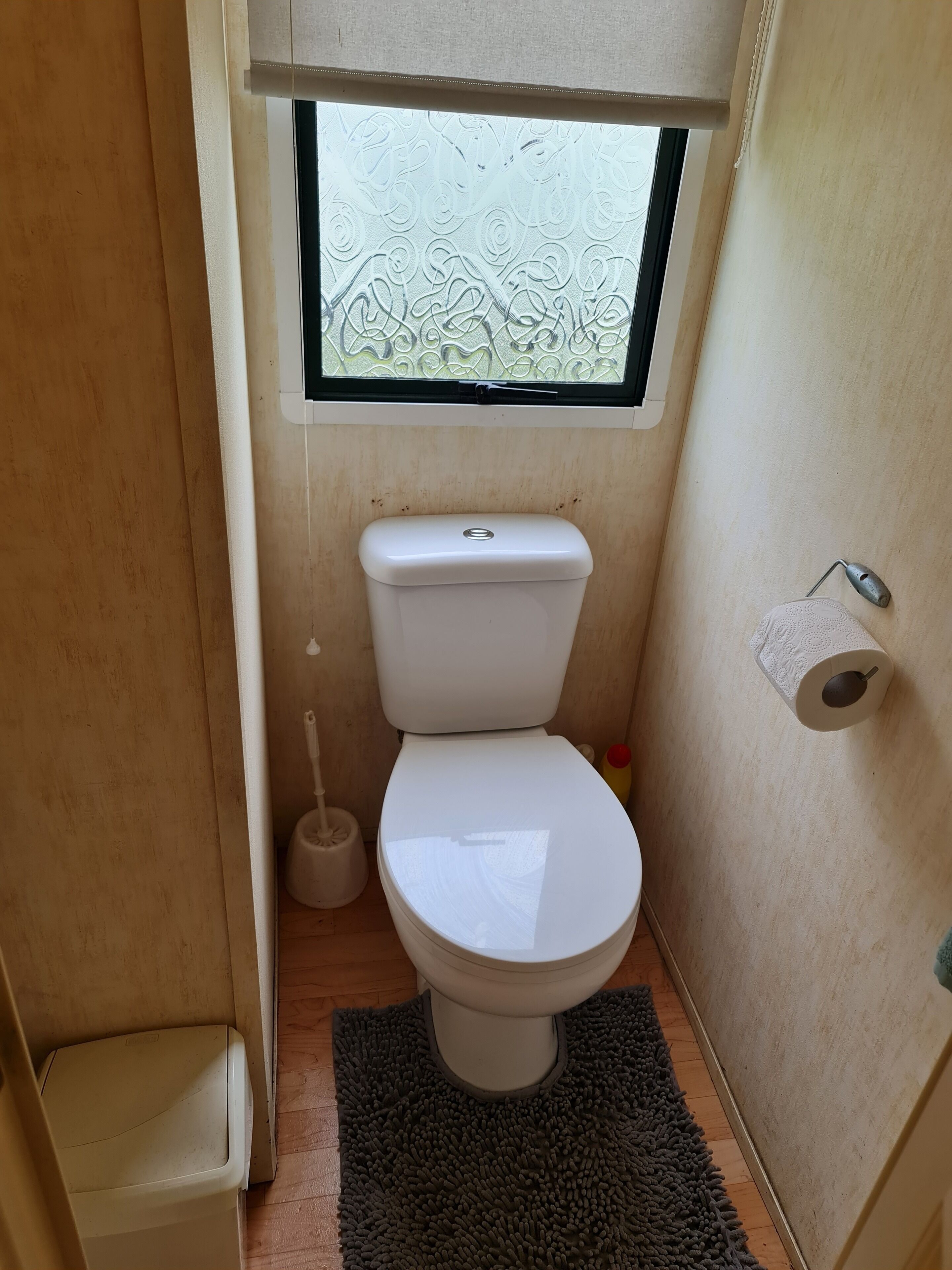 A compact bathroom with a toilet, a window, a toilet paper holder, and a small trash bin.