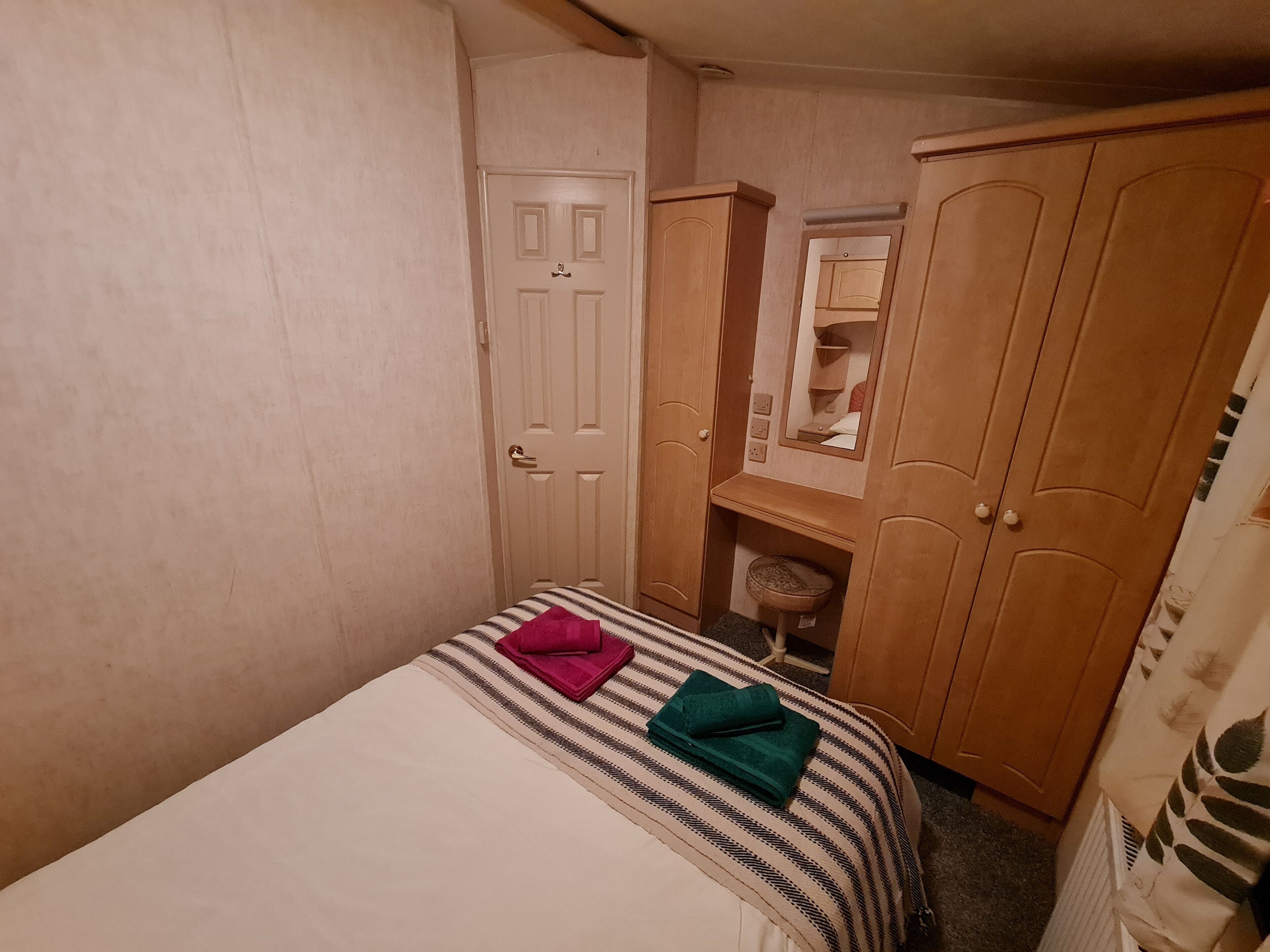 A small, compact bedroom with a single bed, a wooden wardrobe, and a small desk with a mirror.