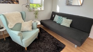 Uma sala de estar com uma poltrona azul-claro, um sofá cinza-escuro, uma mesa redonda pequena com um copo e um tapete preto.