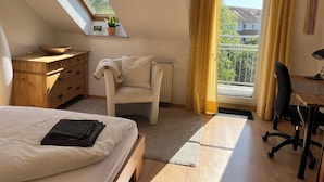 Quarto com cama, escrivaninha, cadeira, cômoda, planta em vaso e janela com cortinas.