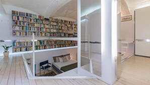 Ein modernes Zimmer mit Schlafplatz in der Schlaffküche, einem großen Bücherregal und einer Glastrennwand.