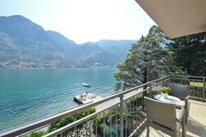 A balcony with wicker furniture overlooking a lake, mountains in the background, and a small boat docked near the shore.