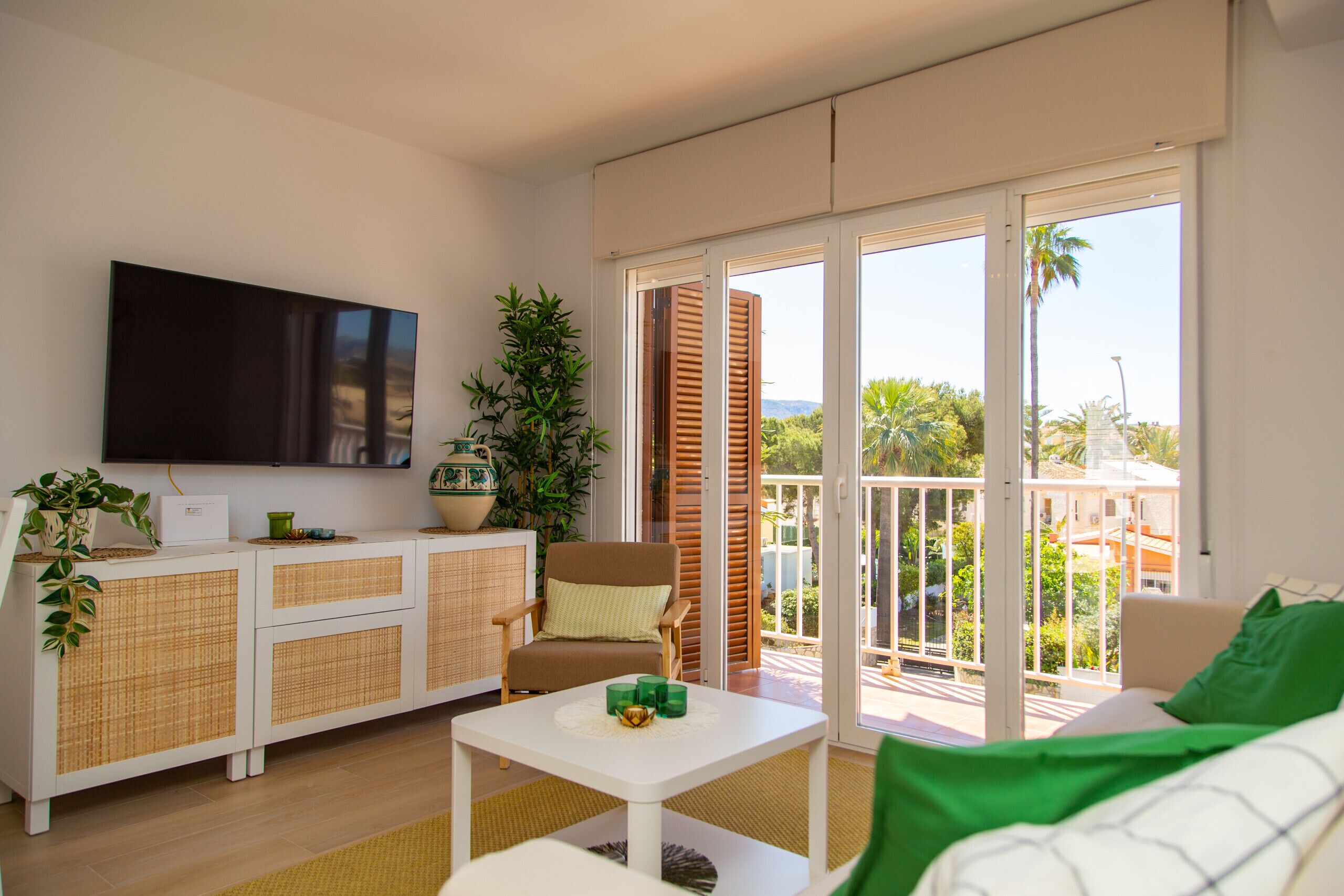 A living room with a flat-screen TV, a white sofa, a small table, and a balcony with a view of palm trees.