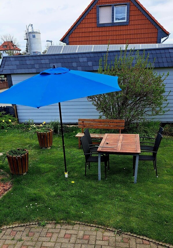 A patio with a wooden table and chairs, a blue umbrella, and a brick pathway.