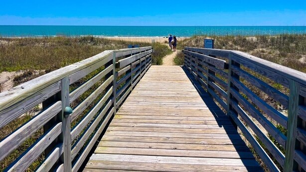 Ein hölzerner Steg führt zu einem Strand unter blauem Himmel.