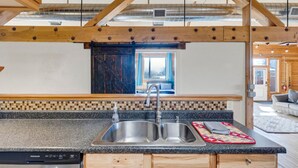 A kitchen with a double sink, wooden beams, a view to the living area, and a tiled backsplash. - The Bitterroot Loft On 1st Street (Hamilton)