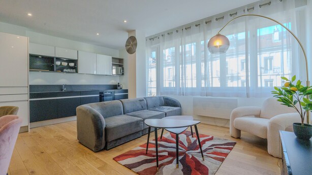 A modern living room with a grey sofa, a small round table, and a red and white rug. There is a kitchen area with white cabinets and a microwave. A large window with sheer curtains allows natural light to fill the space. - #HU | Le Coeur Lorraine 🥐  | City center/Station T3 (Grenoble)