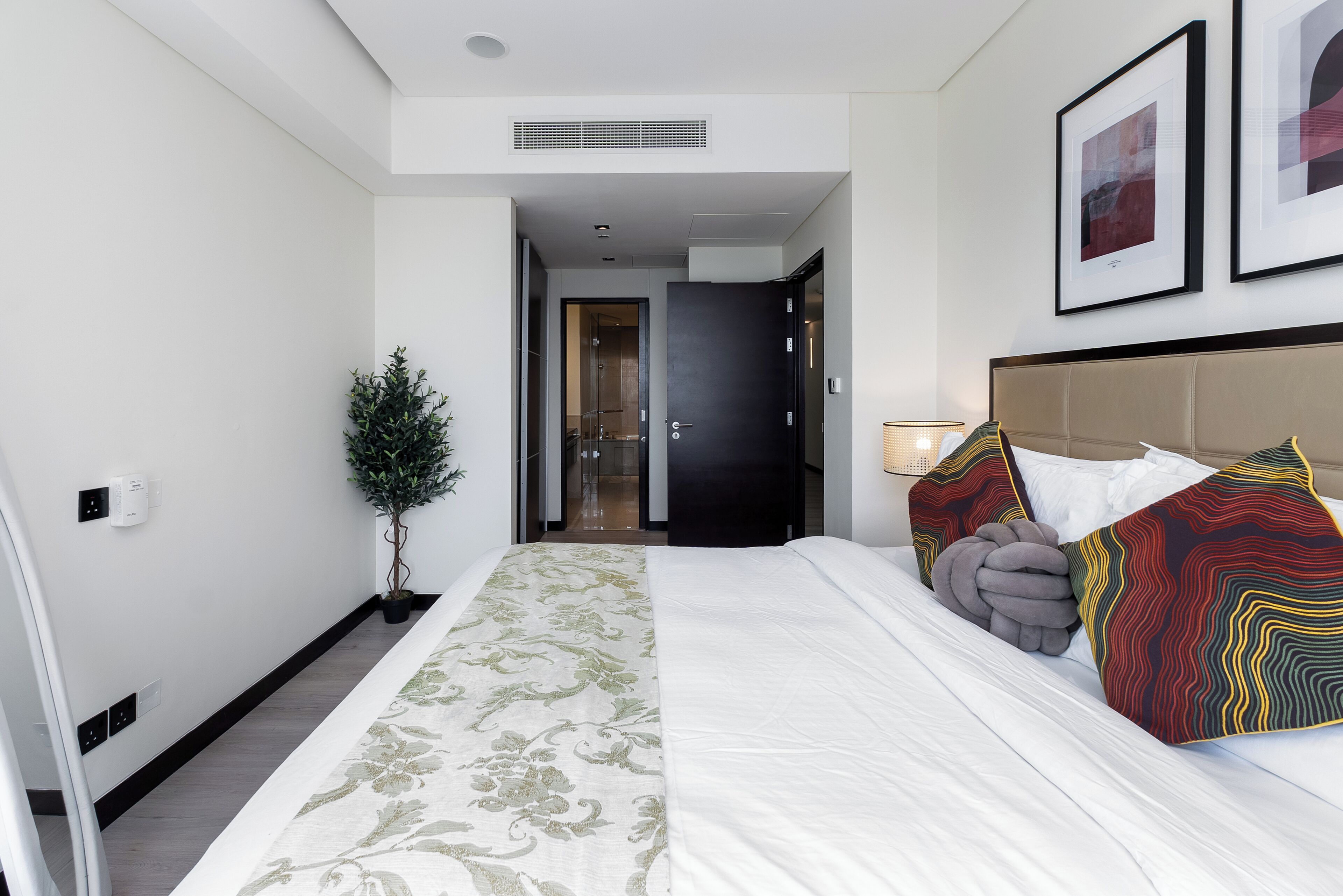 Ein Hotelzimmer mit einem großen Bett, einem Kopfteil, einer Zimmerpflanze und einer Tür zu einem weiteren Raum.