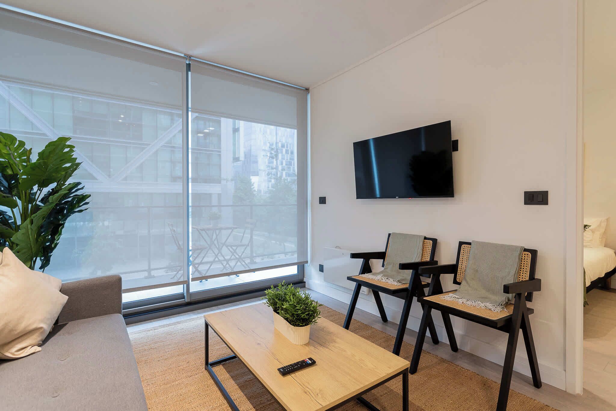 Een moderne woonkamer met een bank, een bijzettafeltje met een plant, en twee stoelen. Een televisie is aan de muur bevestigd.