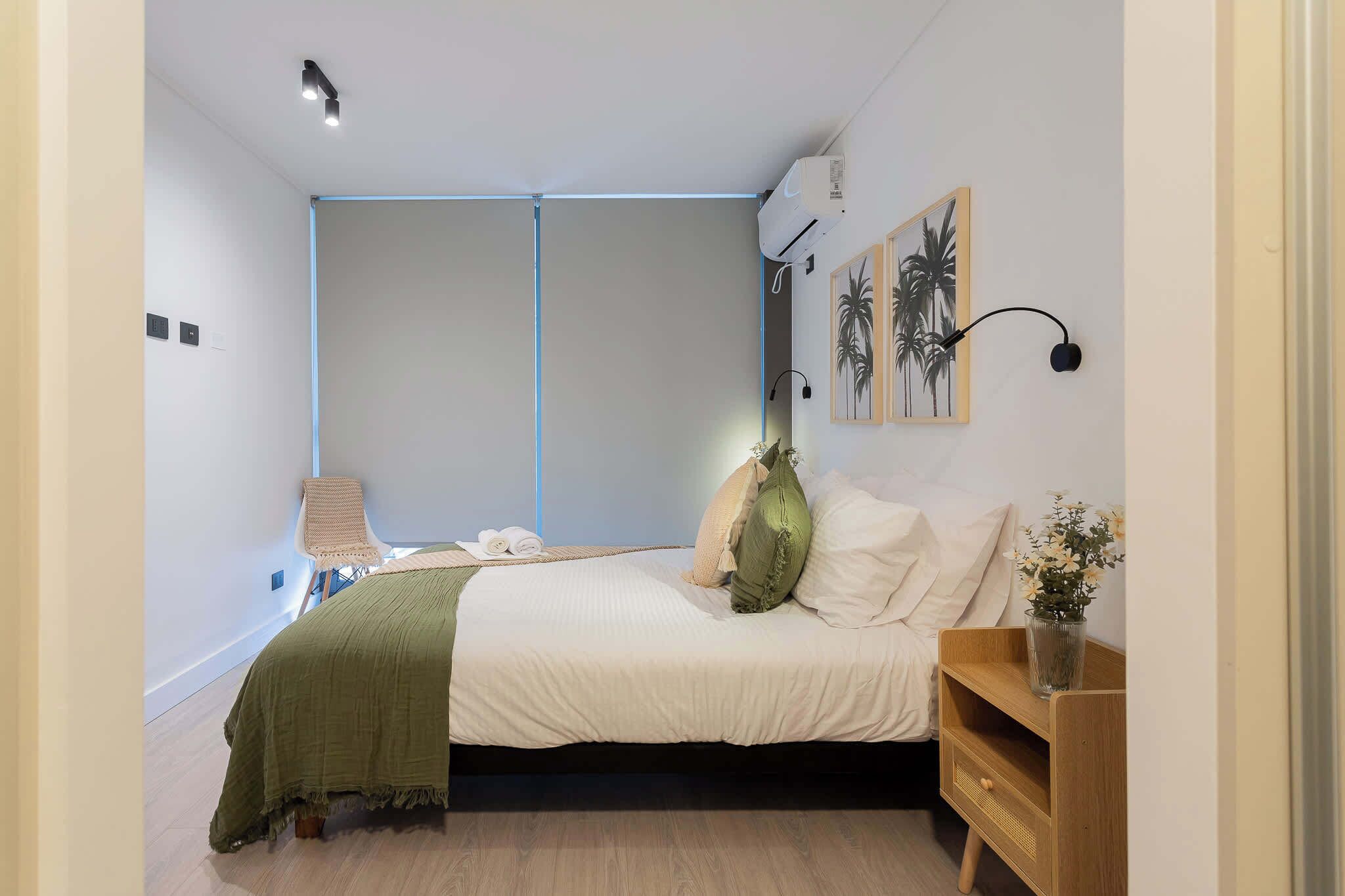 A bedroom with a bed, a bedside table with a vase, a wall-mounted air conditioner, and a painting.