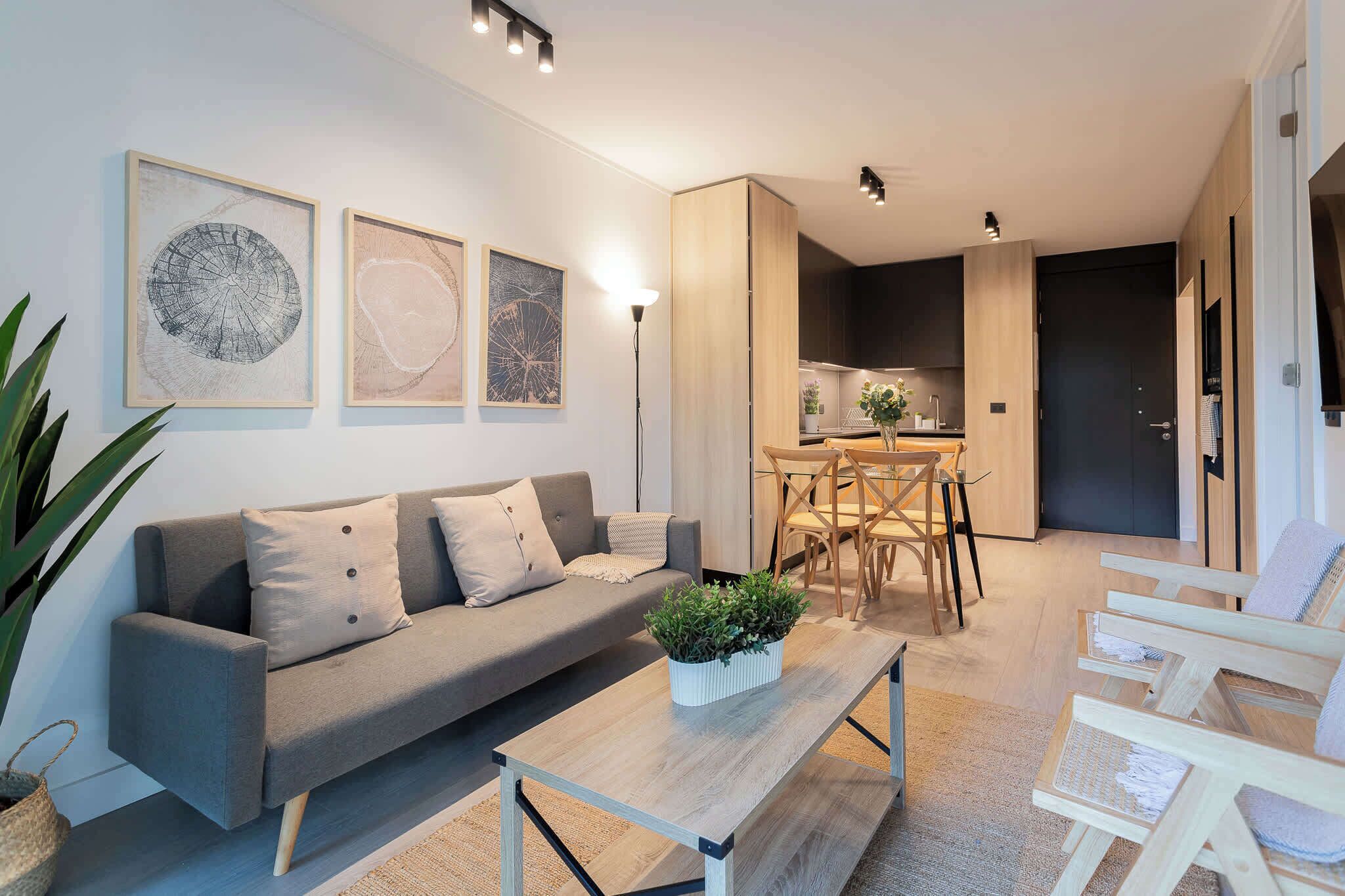 Een moderne woonkamer met een grijze bank, een houten salontafel met een plant en een eethoek met houten stoelen en een tafel.