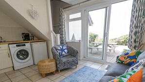 Un espacio habitable compacto con lavadora, un sofá con cojines coloridos, un sillón estampado, un pequeño reposapiés y vistas a un patio al aire libre.