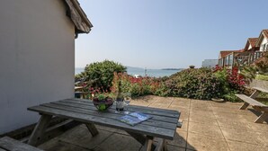 Una mesa de madera con un cuenco de fruta y una botella de vino, frente a un jardín y vistas al mar.
