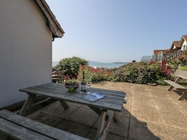 Una mesa de madera con un cuenco de fruta y una botella de vino, frente a un jardín y vistas al mar.