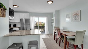 Une cuisine moderne avec des armoires blanches, des appareils en acier inoxydable et un comptoir en granit. On y trouve un coin repas avec une table et des chaises en bois, ainsi qu’un balcon avec vue.
