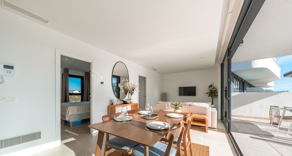 A modern, well-lit room with a dining table set for four, a sofa, a TV, and a large window offering a view of the sea.