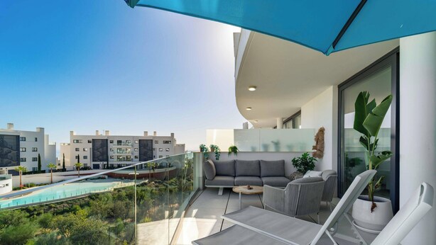 Een balkon met glazen hekwerk, een grijze bank, een witte fauteuil en een kleine tafel.