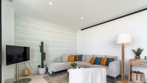 Een moderne woonkamer met een flatscreen-tv, een grijze bank met kleurrijke kussens, een hoge vloerlamp, een cactus en een kleine tafel met een vaas.