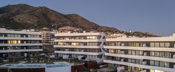 A modern hotel with multiple floors, a pool, and a mountainous backdrop.