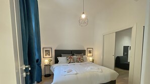 Un dormitorio moderno con una cama grande, dos mesitas de noche con lámparas, una lámpara colgante geométrica y vistas a un pasillo.