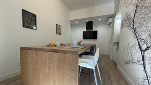 Una cocina moderna con una isla de madera, sillas blancas y una mesa de comedor. Hay un televisor montado en la pared y un altavoz negro.
