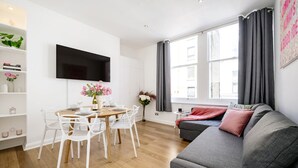 A modern living room with a grey sofa, white chairs, a dining table set for four, and a flat-screen TV mounted on the wall. - Prime West End Flat – Walk to Everything! (Greater London)