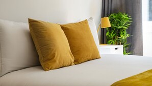A bed with white bedding and two mustard-colored pillows, a bedside table with a lamp, and a green plant in the background. - Prime West End Flat – Walk to Everything! (Greater London)