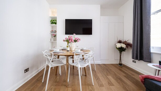 A modern dining area with a round table, white chairs, and a vase of flowers. - Prime West End Flat – Walk to Everything! (Greater London)