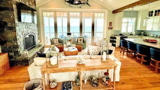 A spacious living area with a stone fireplace, comfortable seating, a wooden coffee table, and large windows offering a view of the ocean.