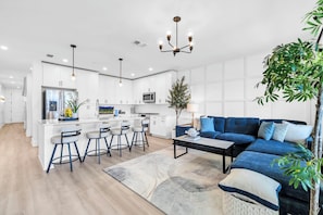 A modern kitchen with white cabinetry, a central island with bar stools, a blue sofa, and a chandelier.