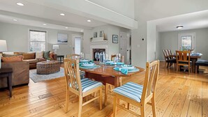 A spacious living room with a wooden dining table set for six, surrounded by wooden chairs with striped cushions. A fireplace with a mantel is visible in the background, and a large window allows natural light to fill the room. - 2 Miles to Coast Guard Beach! (Eastham)