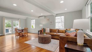 A spacious living room with hardwood floors, a leather sofa, a round ottoman, and a dining area with a table and chairs. - 2 Miles to Coast Guard Beach! (Eastham)