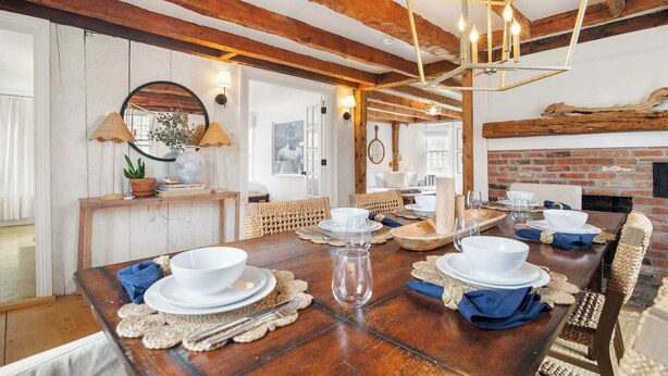 Un comedor rústico con una mesa de madera dispuesta para una comida, una chimenea de ladrillo y vigas de madera a la vista en el techo.